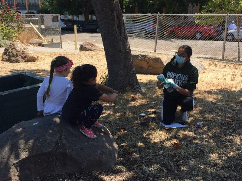 Youth collecting soil samples at an Oakland park while talking to two young children perched on a rock.